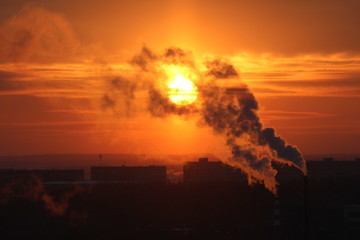 Red sun at sunset or dawn at dusk low above the horizon shrouded in smoke from the chimneys of the plant