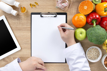 Top view of nutritionist doctor writing meal plan, medical records on clipboard with fresh fruits, vegetables, vitamins, measuring tape and tablet on wooden table. Healthy diet and nutrition concept.