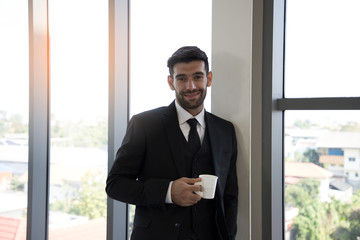 Portrait of a Handsome young businessman with a friendly and smiling broadly at the camera with self-assured expression while holding coffee cup and playing smartphone in the office. Relax time.