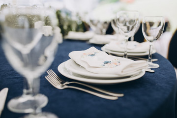 holiday tables are decorated with compositions of flowers and greenery, on the tables are tablecloths, plates with napkins and postcards, glasses, candles and cutlery