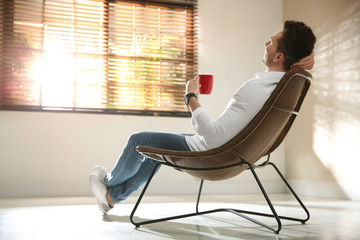 Young man with cup of drink relaxing near window at home. Space for text