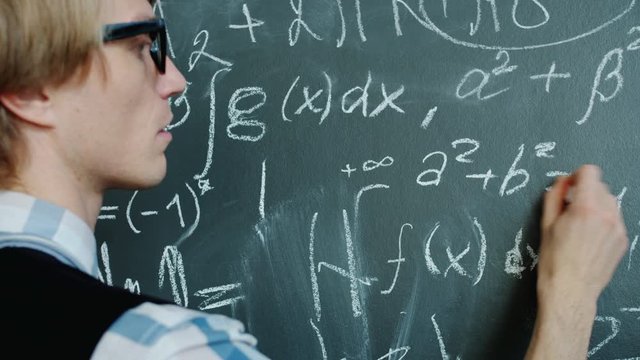 Young mathematician is busy writing formulas on chalkboard working with serious face wearing glasses and casual clothing. People and occupation concept.