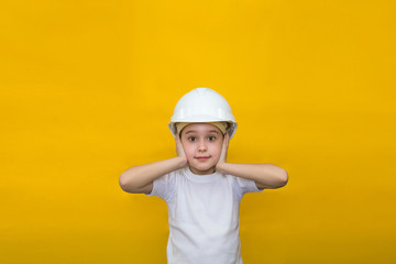 little girl in a construction white helmet covers her ears from loud sounds on a yellow background