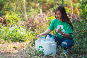 An asian woman making thumbs up hand sign while collecting garbage plastic bottles into a recycle bin in the outdoors