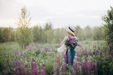 Beautiful romantic woman blonde in a dress and hat in a field with a bouquet of purple flowers of lupins at sunrise. Soft selective focus. Copy space.
