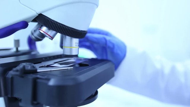 lab technician wearing a white lab coat and blue latex gloves switching magnifying lenses on a microscope 