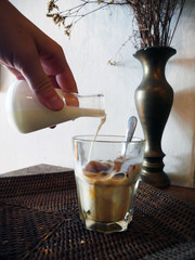 Iced coffee with pouring milk in glass on a wooden background