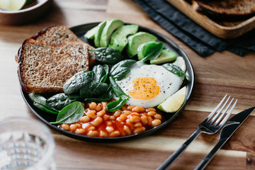 Healthy breakfast or lunch at home or cafe with fried egg, avocado, toasts, beans and fresh spinach on a wooden table.