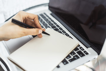 Woman writes with a pen in notebook on laptop keyboard in a sunny office, business and education concept. Close up