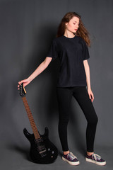 Attractive, young rock girl in black clothes with an electric guitar in her hands stands on a gray background. Model with clean skin.