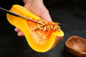 Butternut squash, Woman picks pumpkin seeds. Female hands with steel knife, wooden bowl on black...