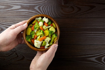 Vegetable mix. Vegan food. Boiled broccoli, cauliflower and carrot in wooden bowl in female hands on wooden background, top view. Healthy food. Steamed vegetables