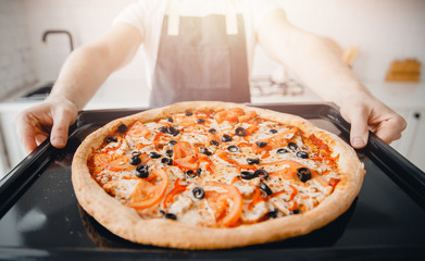 Hot fresh Italian pizza on baking sheet from oven, chef in apron holds in hands, sunlight