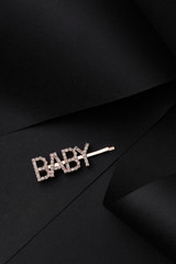 Subject shot of a pink gold hair clasp made as a sign plate with sparkling crystal lettering "BABY". The sparkling hairpin is isolated on the black silky surface. 