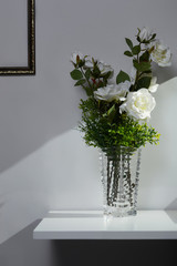 Subject shot of a cut-glass vase with geometric design and with a bunch of white roses and green leaves in it. The vase is located on the white table near the gray wall with a bronze picture frame. 
