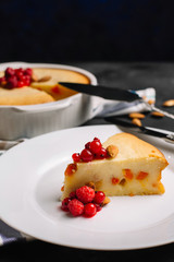 Freshly baked homemade cottage cheese pie with dried apricots, almonds and raspberries. A piece on a white plate decorated with berries and Cutlery side View on a dark background