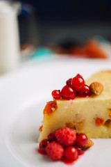 Freshly baked homemade cake with cottage cheese and dried apricots, almonds and raspberries and a glass of milk. A piece on a white plate decorated with berries and Cutlery side View on a dark backgro