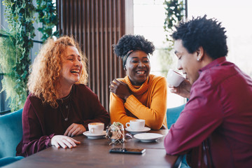 Three friends sitting in a cafe, talking and laughing. There are two young ladies and a guy.