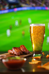 Spicy barbecue chicken wings, tomato salsa, nachos and beer on dark wooden bar table. Football on...