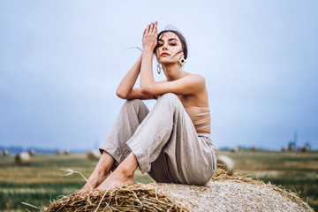 Brunette in linen pants and bare shoulders sitting on a hay bales in warm autumn day. Woman looking at camera. Behind her is a wheat field