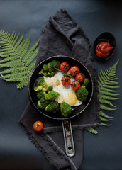 Healthy breakfast: broccoli, cherry tomatoes and fried eggs in a pan, which stands on a wooden stand on a dark background. 