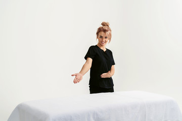 Portrait of young smiling friendly funny woman masseur in uniforme near massage table in Spa Salon. Physical Therapist In Medical Office. Beauty Treatment, Massage Therapy. White background isolated - obrazy, fototapety, plakaty