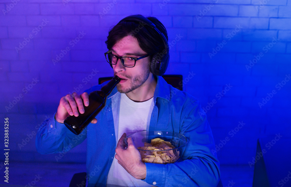 Wall mural millennial guy playing online game drinking beer at home, low-light