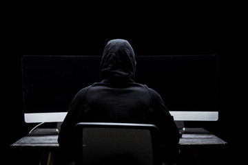 back view of hacker sitting near computer monitors with blank screen isolated on black