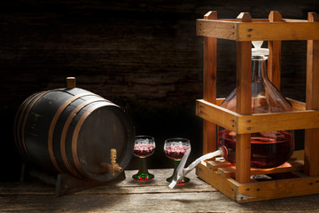 Wine balloon with red wine in a wooden rack, glasses and barrel in the wine cellar