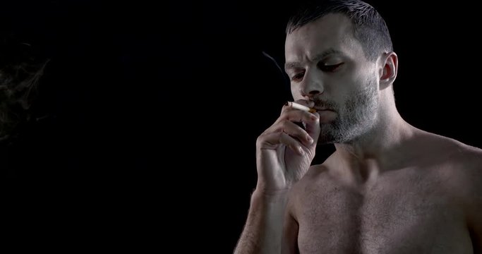 portrait of an unshaven brunette with very pale skin on a black background. he smokes a cigarette, takes a drag, opens his mouth, lets out a lot of smoke