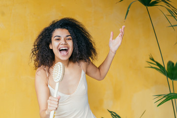 Recycling, zero waste, eco friendly concept. African American mixed race woman dressing beige bathrobe with bowl and singing holding body brush as a microphone on yellow background.