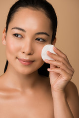 smiling beautiful naked asian girl holding cotton pad on face isolated on beige