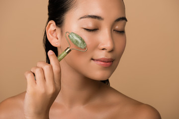 beautiful naked asian girl with closed eyes massaging face with jade roller isolated on beige