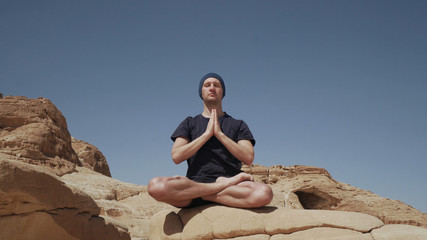 Naklejka na ściany i meble Attractive male sitting in lotus pose and doing namaste on a rock in desert