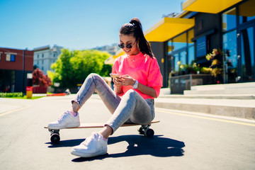 Happy female skater sitting at girlish longboard and using cellphone device for watching video of skating tricks, youthful woman in sunglasses recreating in city connecting to 4g for chatting