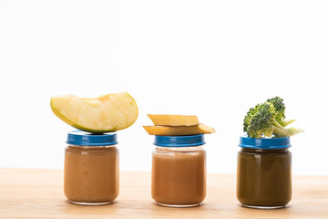 Jars of baby food with pieces of apple, broccoli and banana slices on wooden surface isolated on white