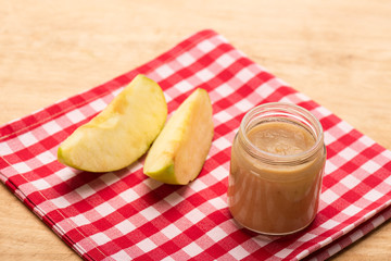 Jar of fruit puree with apple pieces on checkered napkin on wooden surface