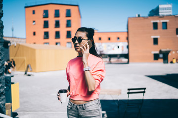 Asian female student in trendy sunglasses standing at urban street and making international conversation during coffee break,attractive millennial woman phoning via cellular application and roaming 4g