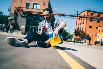Caucasian male blogger with education textbooks in hand sitting at asphalt road in campus and...