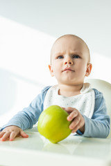 Fototapeta premium Selective focus of pensive baby boy holding green apple while sitting on feeding chair on white background