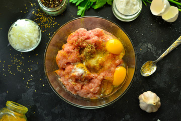 Cooking process. Meat, eggs, spices, parsley,  onions in a glass bowl. Ingredients for Meatballs. Top view. Food and ingredients on a dark table background. Minced chicken.