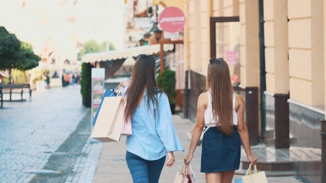 Two beautiful girls have fun walking down the street with their purchases after shopping. Back view. Copy space. 4K.