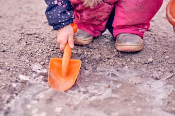 Low section of a 4 year old child girl in muddy dirty pink winter trousers squatting and working on the ice on a frozen puddle with an orange  shovel. Seen in Germany in February.