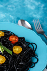 closeup unusual dish of black spaghetti with cuttlefish ink with yellow and red cherry tomatoes in blue plate on blue background with fork and spoon