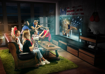 Group of friends watching TV, match, championship, sport games. Emotional men and women cheering for favourite MMA fighter of America with flag. Concept of friendship, sport, competition, emotions.