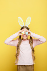 cheerful child with bunny ears covering eyes with easter eggs isolated on yellow