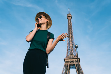 Excited Caucasian female traveller enjoying smartphone conversation with friend during holidays time for visit romantic showplace, joyful woman in sunglasses and hat calling via mobile network app