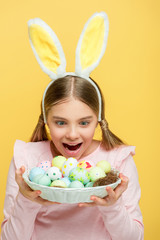 excited child with bunny ears looking at easter eggs in basket isolated on yellow
