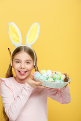 cheerful kid with bunny ears sticking out tongue and holding easter eggs in basket isolated on yellow