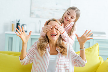 happy kid covering eyes of surprised mother gesturing in living room
